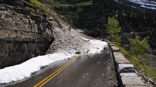 Snow piles onto a mountain road blocking traffic with trees in the background. 