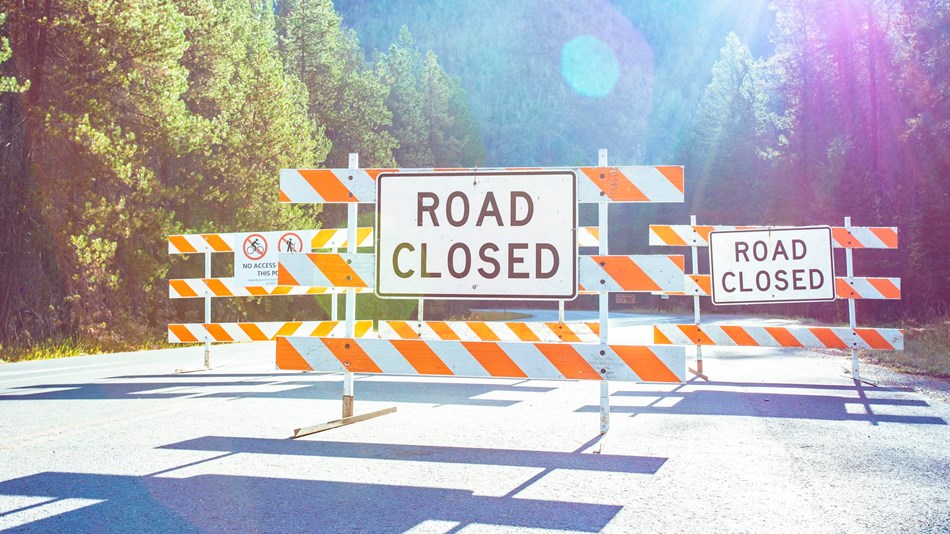 A road is closed with a large sign with the sun flaring behind.