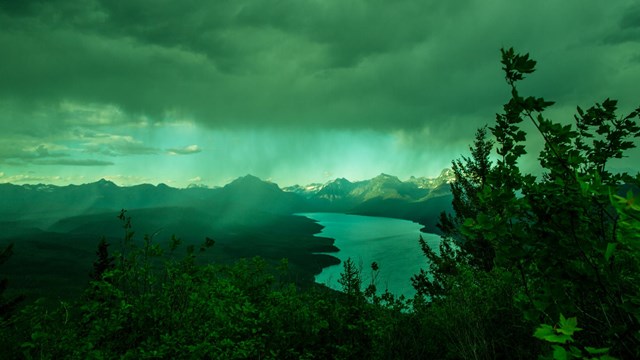 A bolt of sunlight shines through cloudy and hazy skies, hitting mountains and a lake below.
