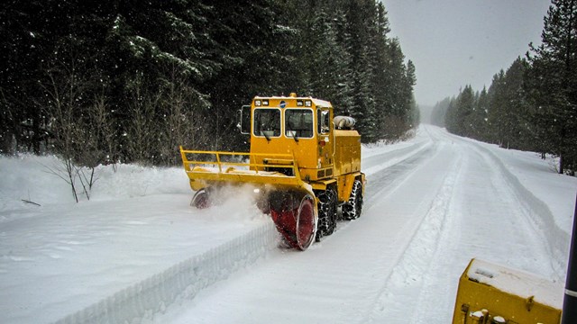 A large yellow snow plowing machine moves snow off a high mountain road. 