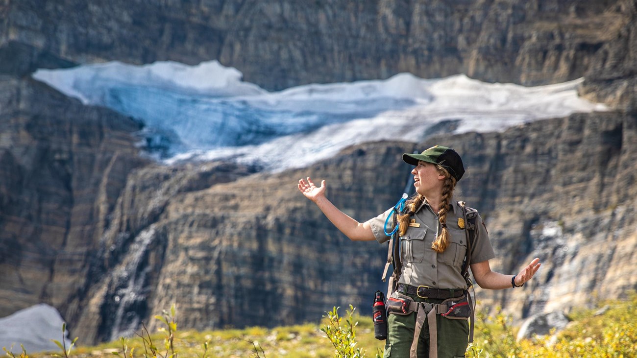 A park ranger stands with their arms out in front of a glacier. 