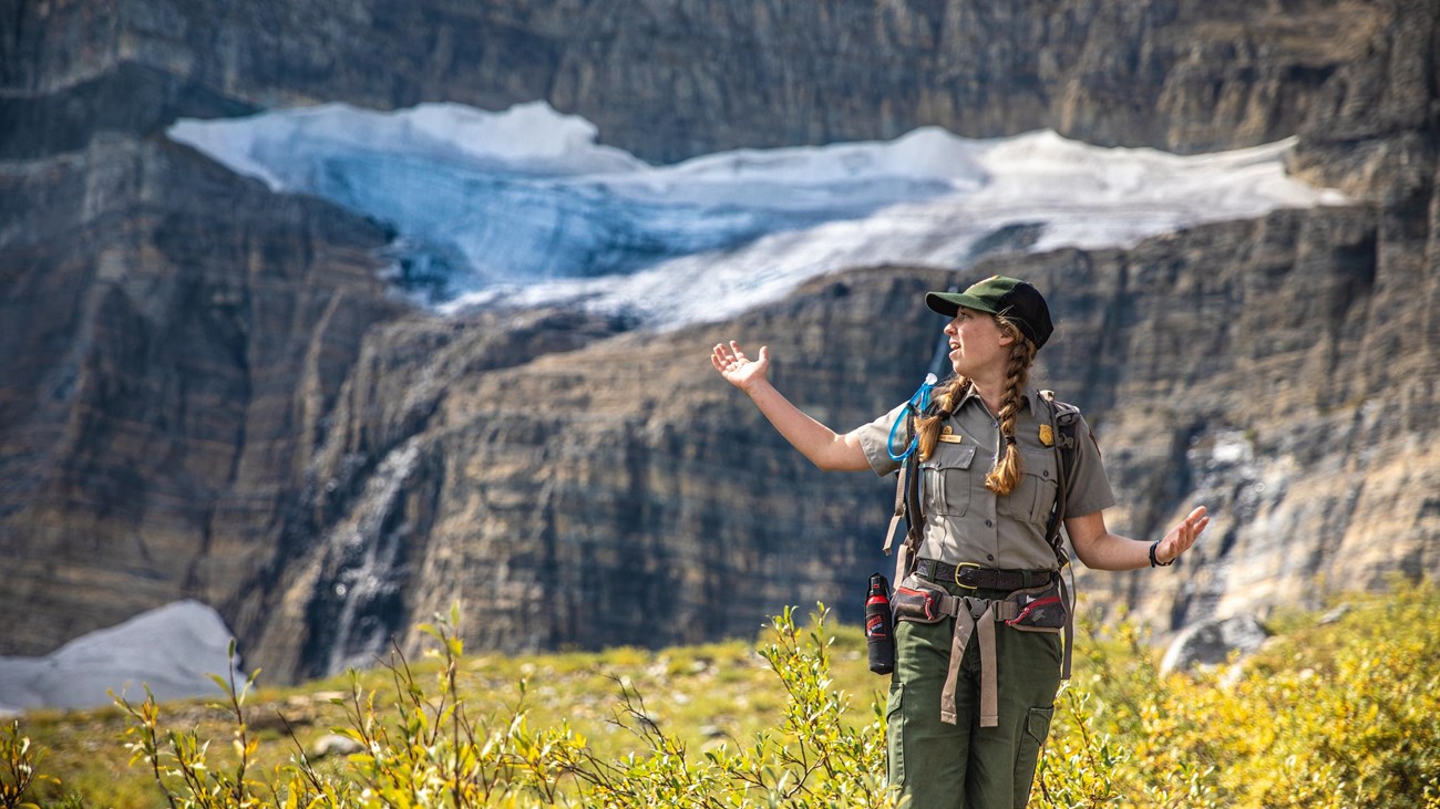 A park ranger stands with their arms out in front of a glacier. 