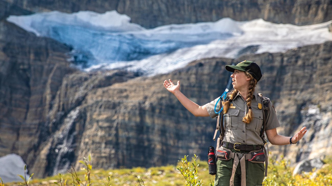 A park ranger stands with their arms out in front of a glacier. 