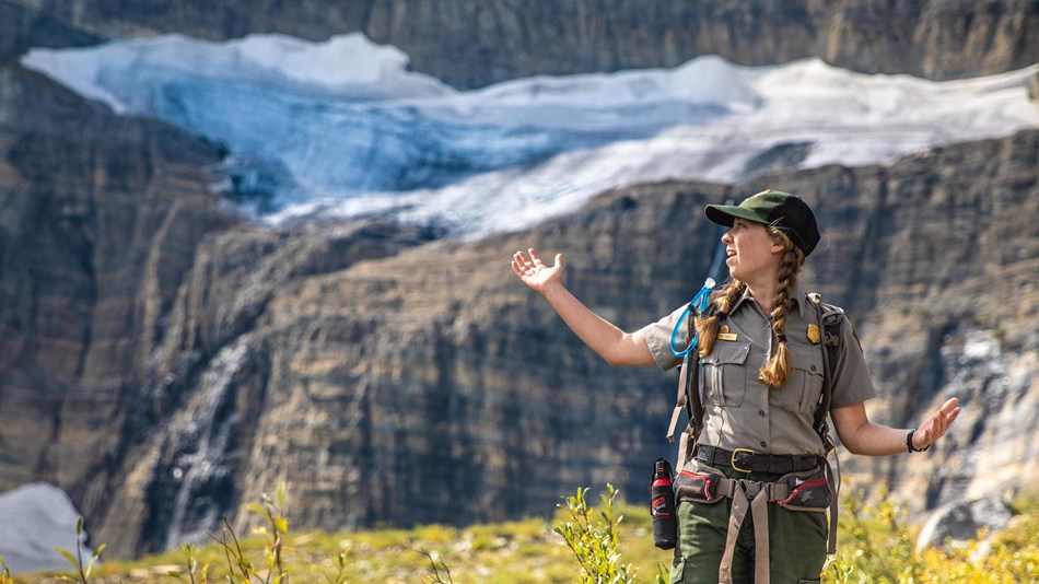 A park ranger stands with their arms out in front of a glacier. 