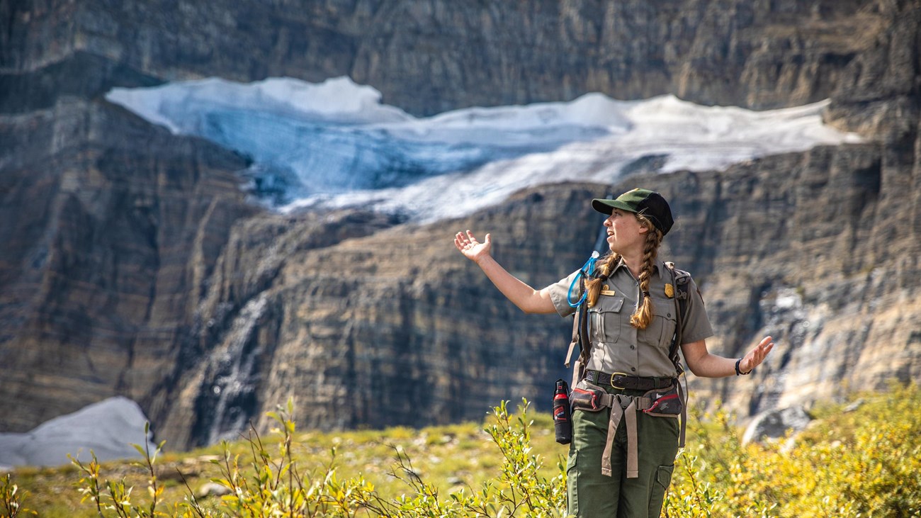 A park ranger stands with their arms out in front of a glacier. 