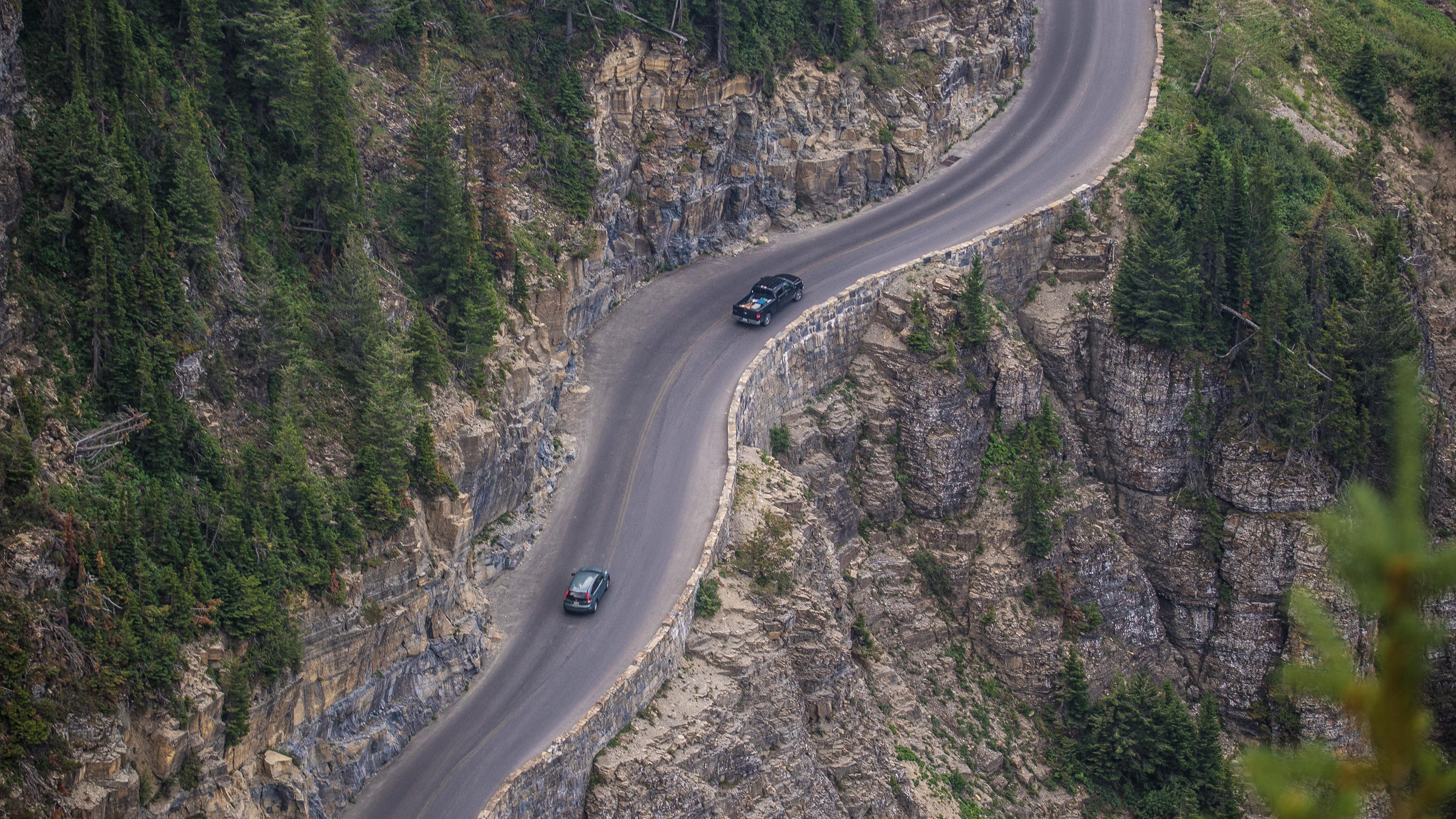 Two cars drive along a cliff's edge narrow road with trees all around. 