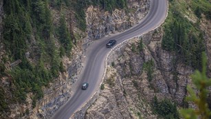 Two cars drive along a cliff's edge narrow road with trees all around. 