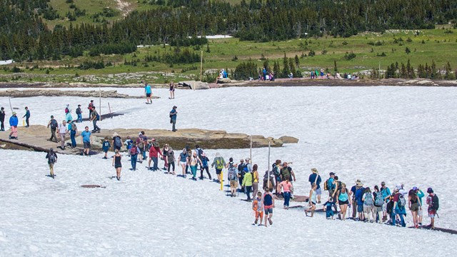 Crowds of people walk over a snowy trail in an alpine area. 