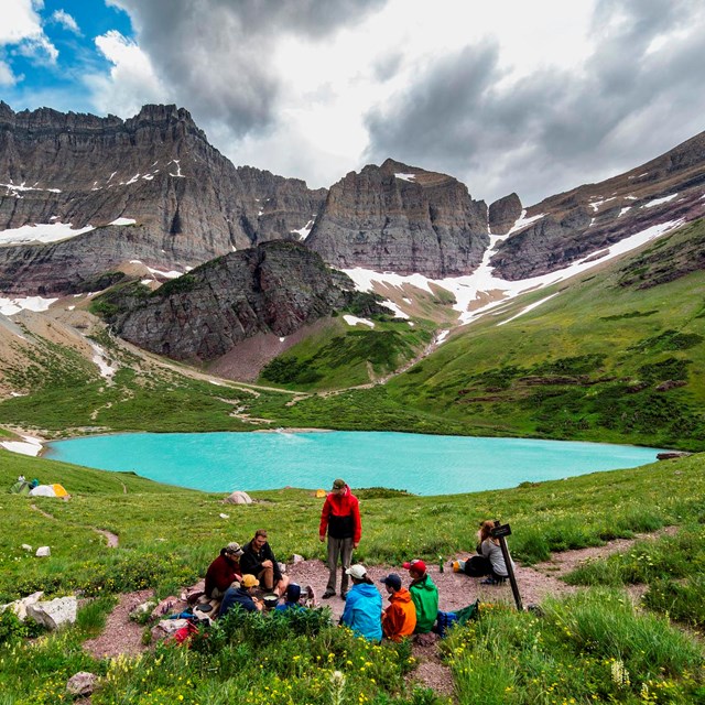 Visitors prepare a meal at a lakeside wilderness campsite near a mountain.