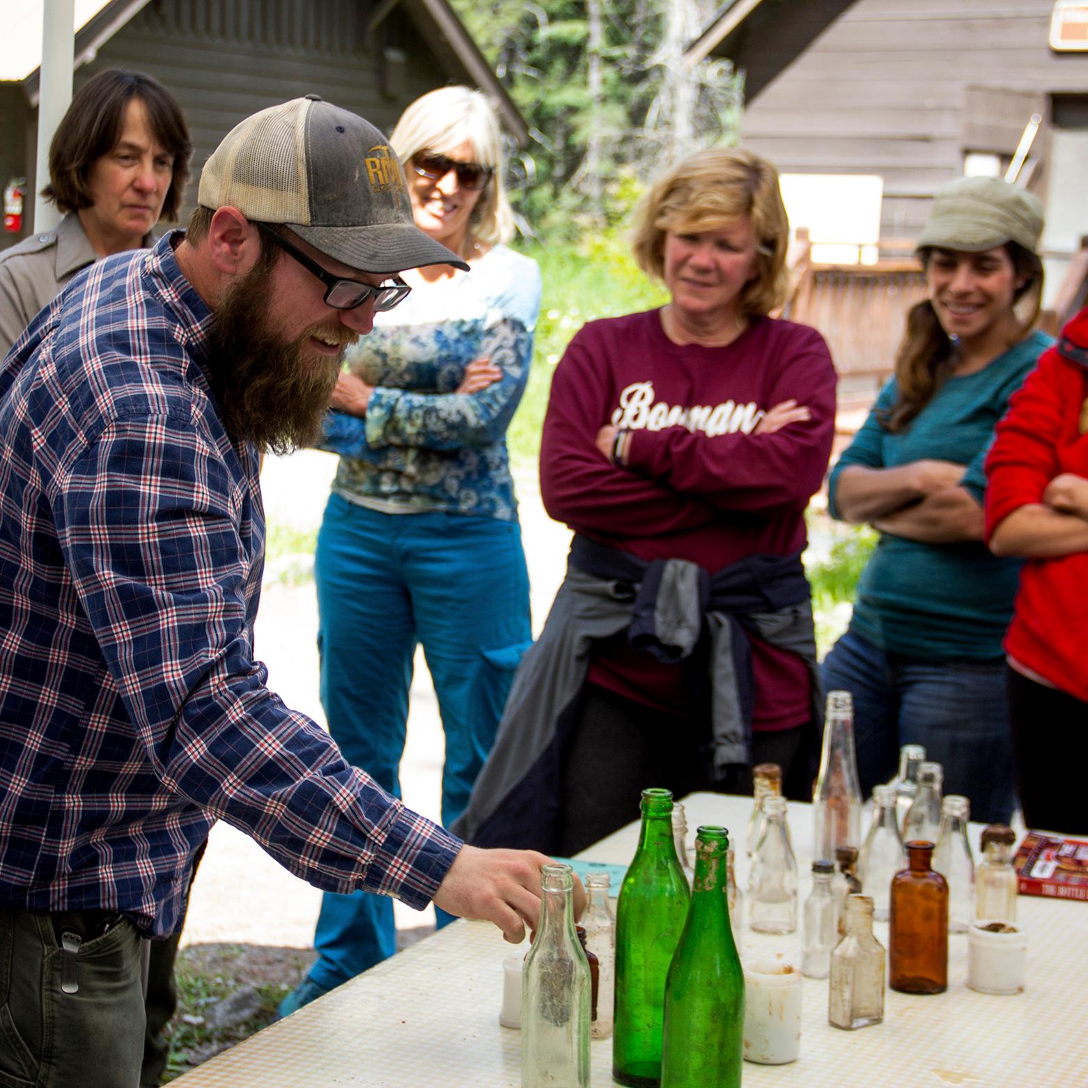 Group of teachers gather around table where bearded man points to different glass bottles