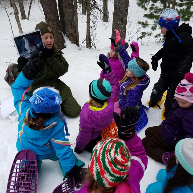 All seated in snow, children wearing coats and snowshoes raise their hands in front of a ranger