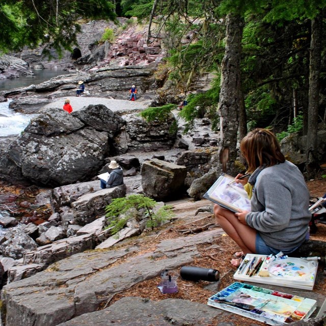 Woman sits by stream and paints with watercolor, other painters in background