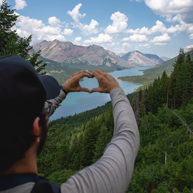 A hiker holds his hands in a heart before a lake and mountains.