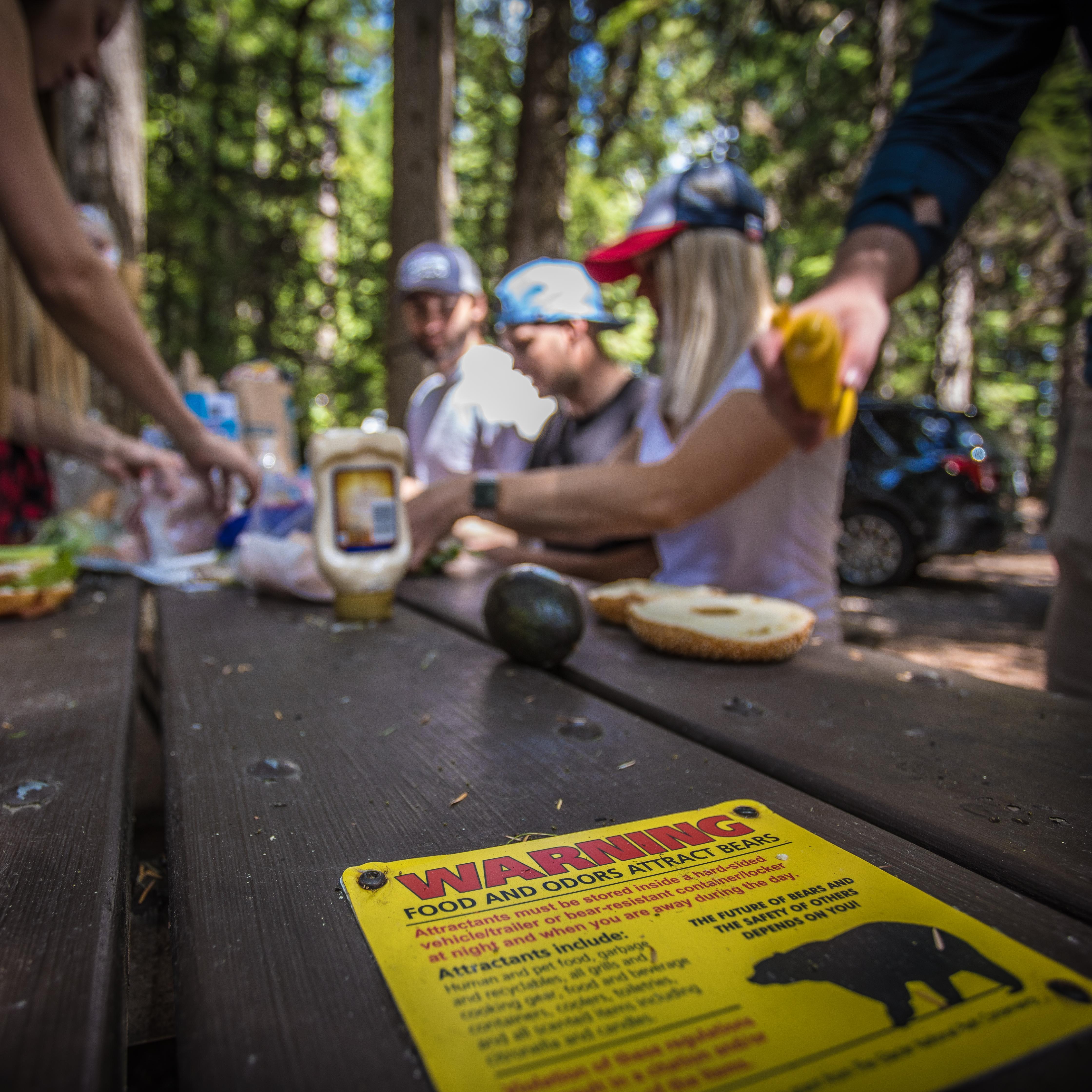 A group sits in a campground preparing a meal, in the foreground a bear safety sign.
