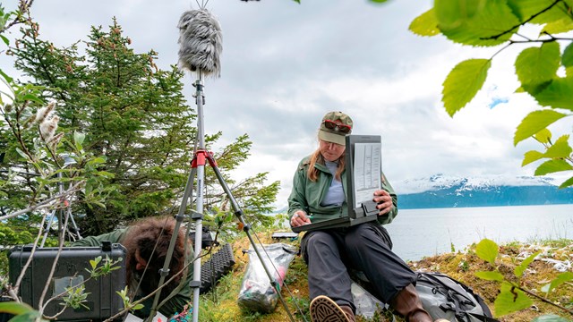 Two researchers operate a hydraulic pulley on a research vessel in front of a glacier.