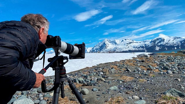 A man looks through a camera at a scene with rocks, snow, mountains, and blue sky. 