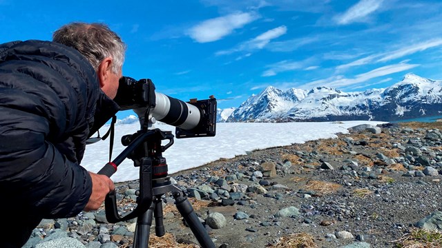 A man looks through a camera at a scene with rocks, snow, mountains, and blue sky. 