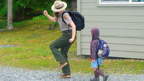 a small child walks behind a park ranger on a path near a building