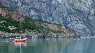 A sailboat sails through calm water with steep mountain cliffs behind