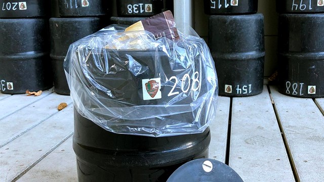 A black, cylindrical bear-resistant food canister with a clear plastic liner and some packaged food