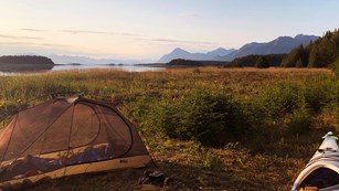 a tent and a kayak sit on an island, surrounded by water, mountains, and a sunset sky