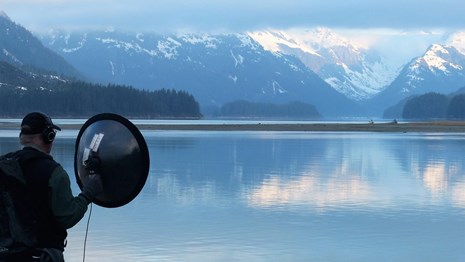 a person stands with sound measuring equipment near a lake and mountains