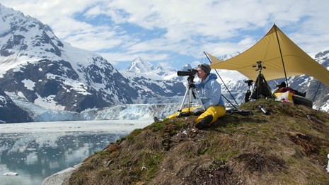 researchers sit under a tent looking through binoculars near a tidewater glacier