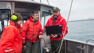 three people in bright coats on board a ship look at a computer