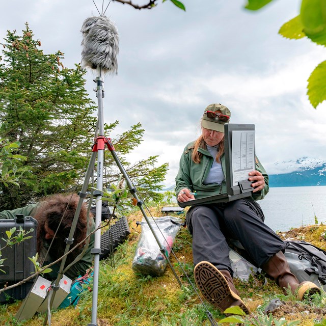 Two researchers operate a hydraulic pulley on a research vessel in front of a glacier.