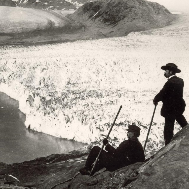 Two people look down at a huge tidewater glacier from a mountainside.