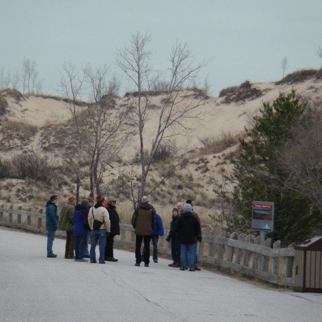 A group of people listen to a talk while standing on a sidewalk at the foot of a tall sand dune.