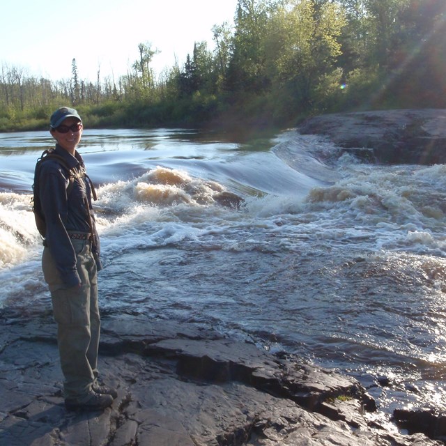 A woman stands on flat bedrock as a stream tumbles behind and past her.