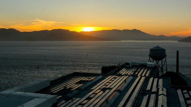 sun setting over a body of water and mountains, water tank and roof with solar panels in foreground