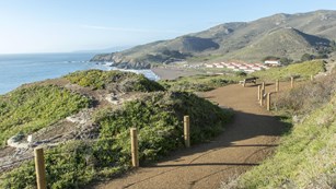 A trail runs across the hills overlooking a beach and red roofed buildings.