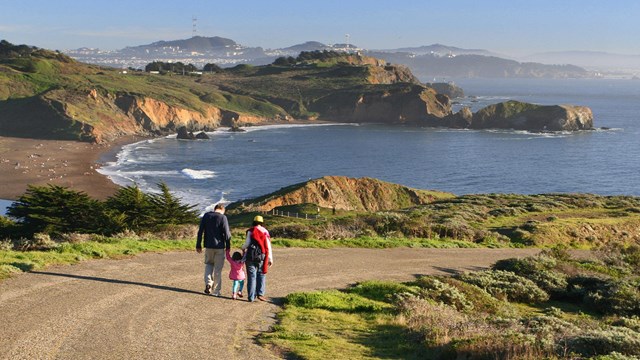 A couple with small child walk down a road with Rodeo Beach, ocean, Bird Rock and city behind