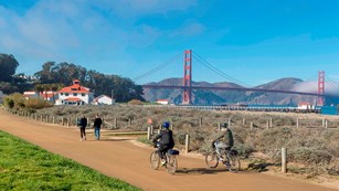 Two cyclists bike and two individuals walk along a path in front of a large red suspension bridge