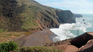 Waves crash against a sandy cove surrounded by chaparral covered bluffs. 