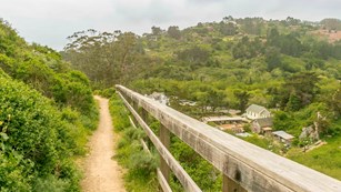 A trail runs between a chaparal covered hillside and a wooden fence. It overlooks the valley below. 