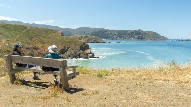 Two individuals sit on a wooden bench overlooking coastal bluffs and the Pacific ocean. 