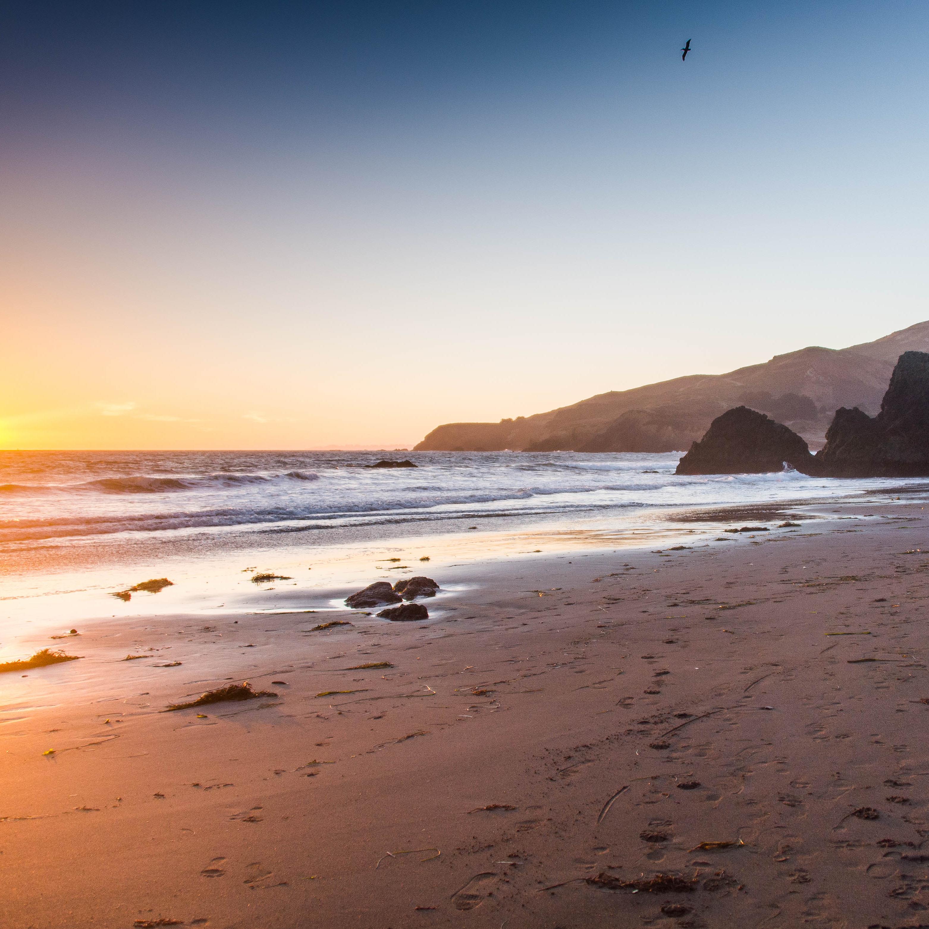 The sun sets on a beach with large rock formations. Short buildings are visible in the background.