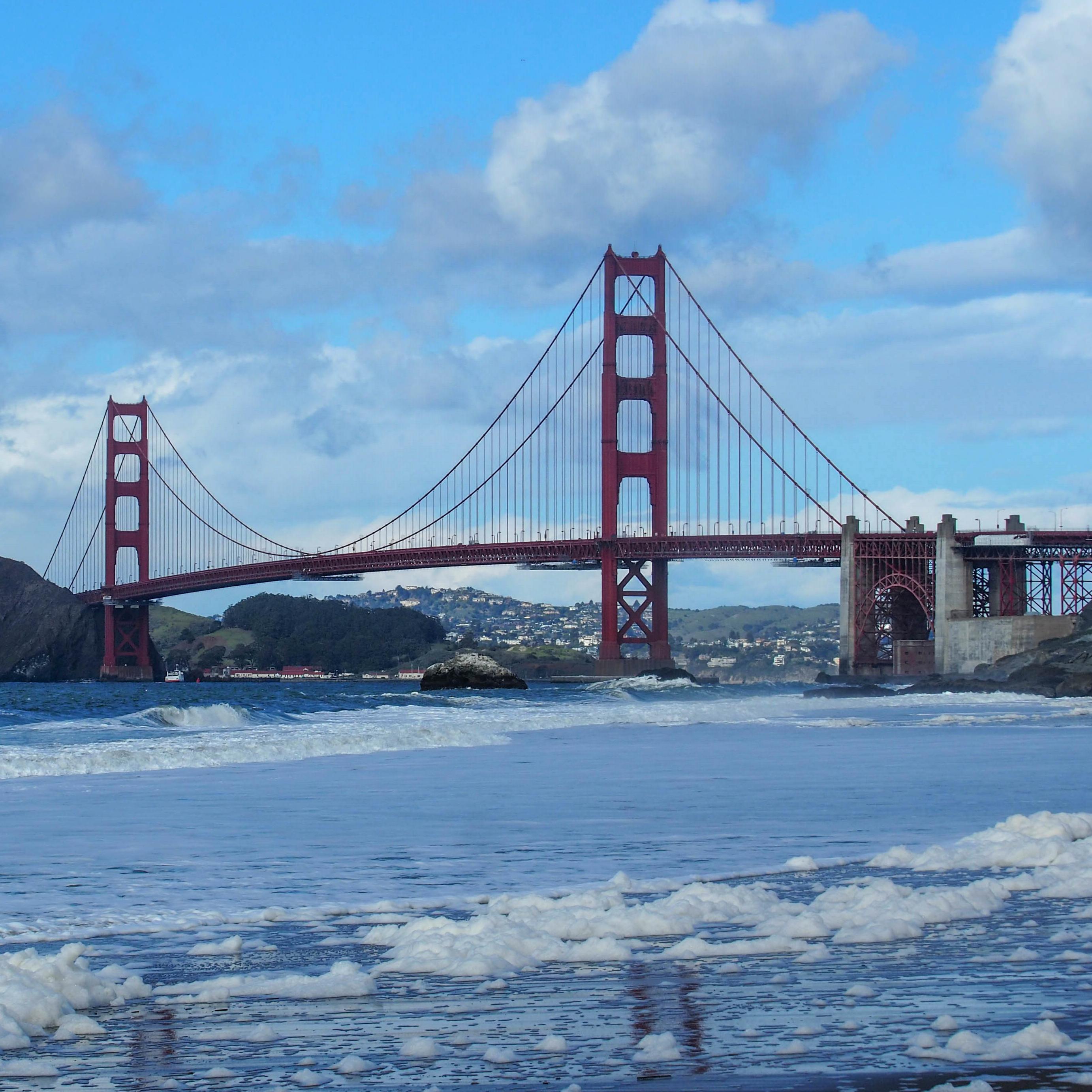 Sea foam covers a beach with fort point and the golden gate bridge in the background.