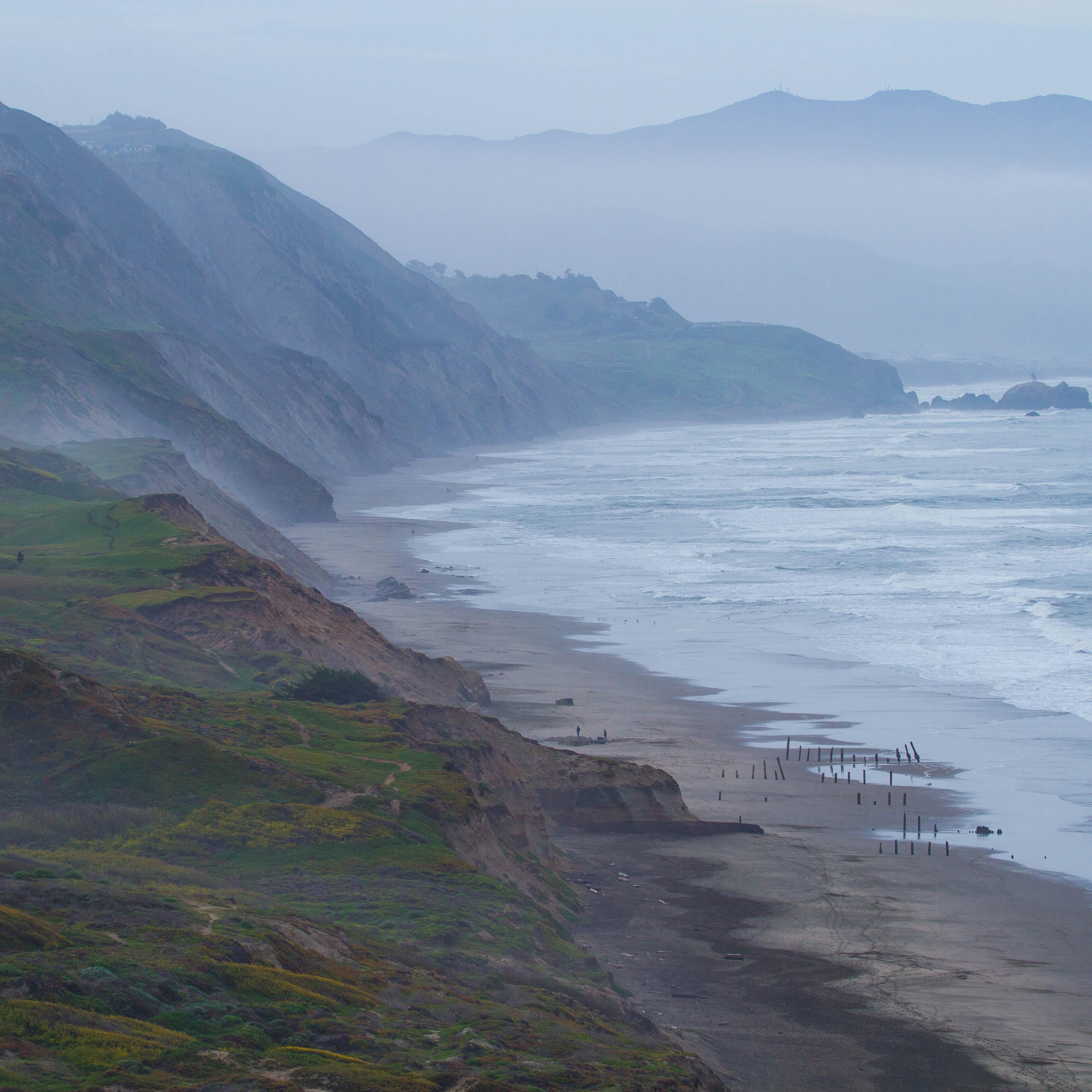 Waves crash on the shore surrounded by rolling green hills on a foggy day.