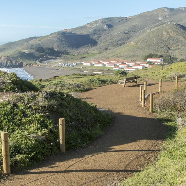 A trail runs across the hills overlooking a beach and red roofed buildings.