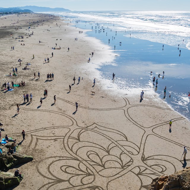Ocean beach crowds on a beautiful day, including someone creating sand art with a rake
