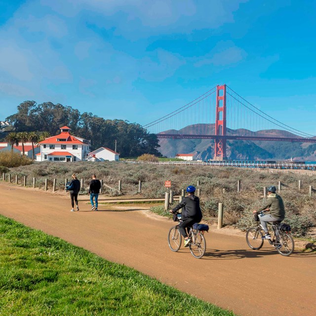 Two cyclists bike and two individuals walk along a path in front of a large red suspension bridge