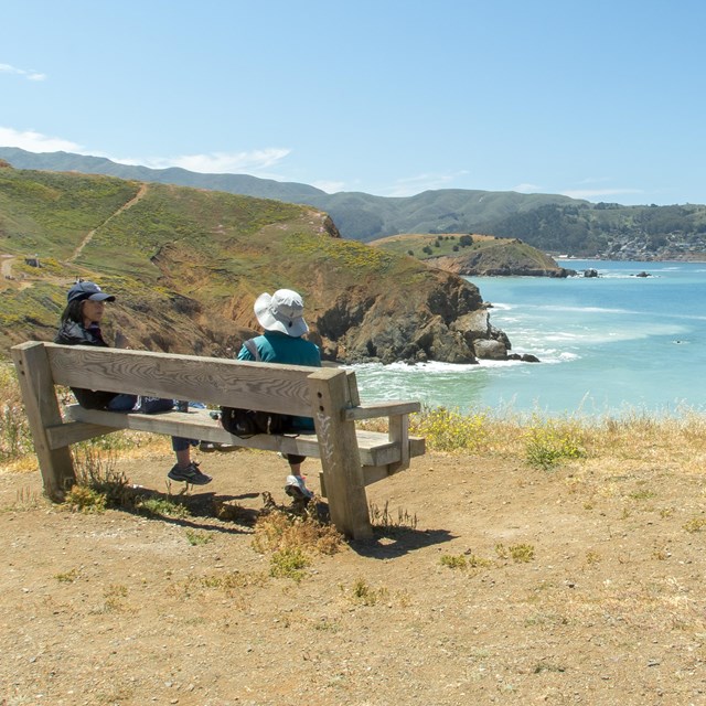 Two individuals sit on a wooden bench overlooking coastal bluffs and the Pacific ocean. 