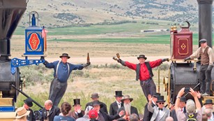 People in period clothing stand next to the steam locomotives.
