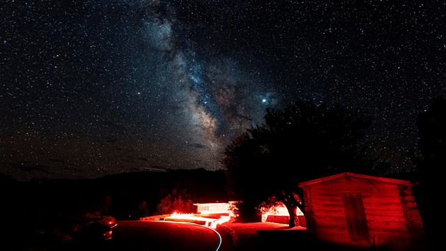 Milky Way over a red-lit Lehman Caves Visitor Center, with a historic cabin in foreground.