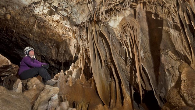 A caver sits on a rock with a headlamp illuminating draperies clinign to a rock wall.