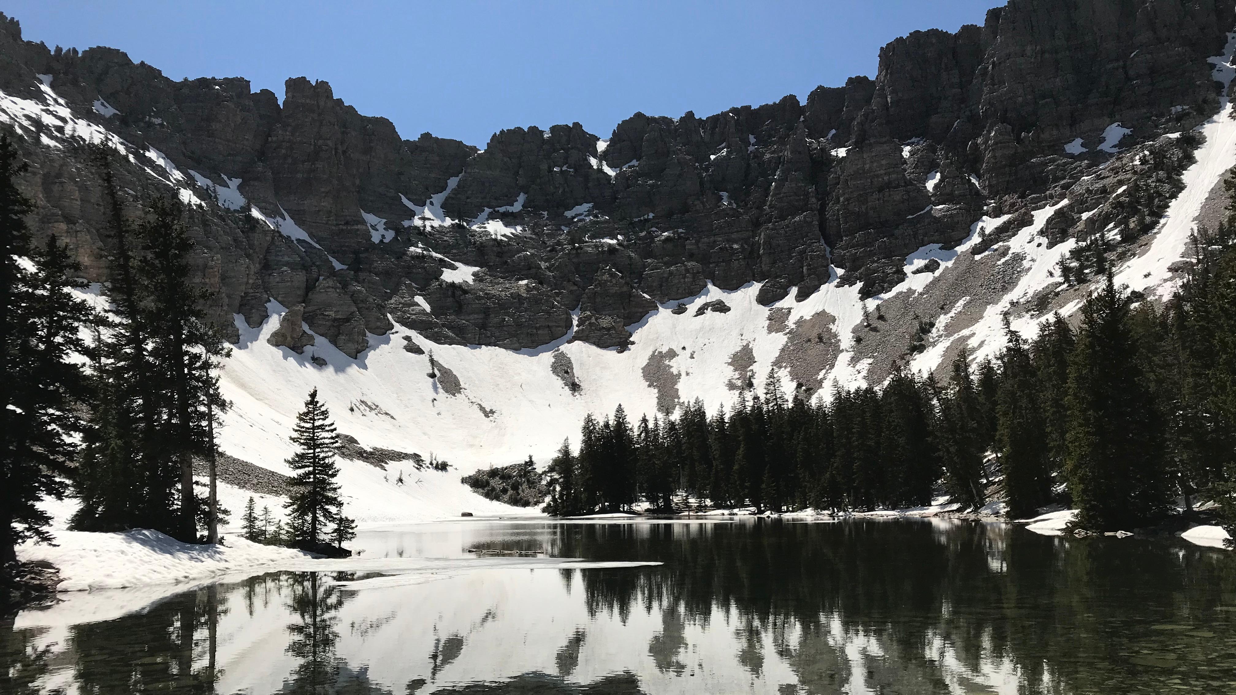 An alpine lake surrounded by tall pine trees is overlooked by sheer grey cliffs with patchy snow